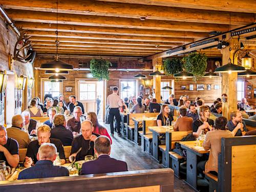 A busy wooden restaurant interior with many people dining at tables and a server walking between them. The space is warmly lit and decorated with plants and wall-mounted antlers.