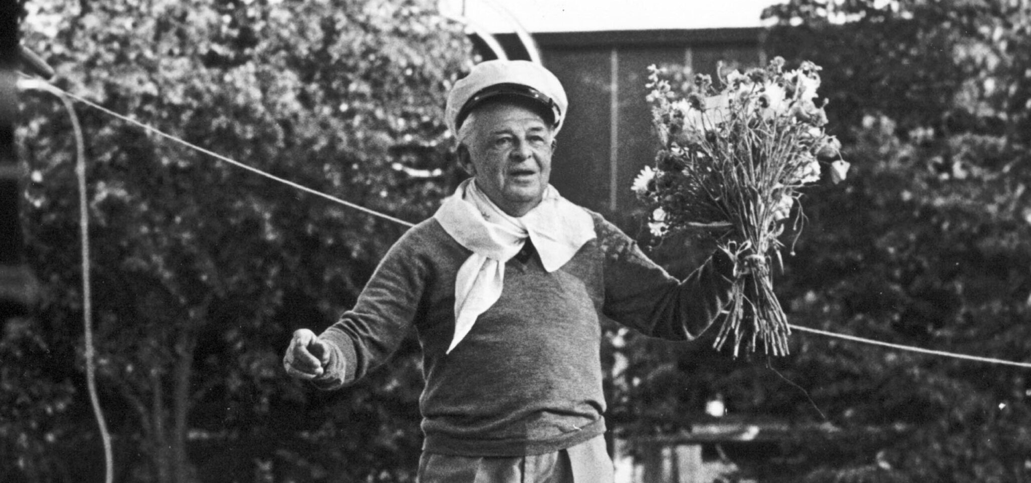 An older person wearing a sweater, scarf, and cap holds a bouquet of flowers outdoors, standing near a rope with trees in the background.
