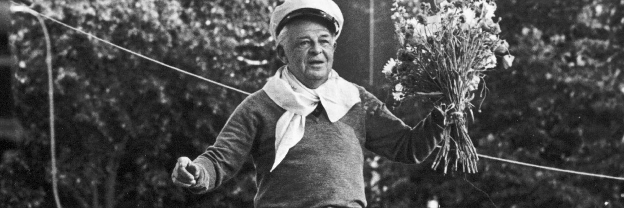 An older person wearing a sweater, scarf, and cap holds a bouquet of flowers outdoors, standing near a rope with trees in the background.