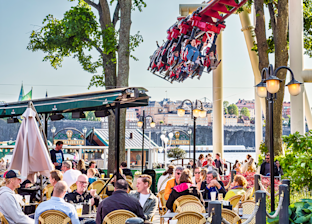 Människor sitter vid cafébord utomhus medan en berg- och dalbana med passagerare passerar ovanför i en nöjespark.