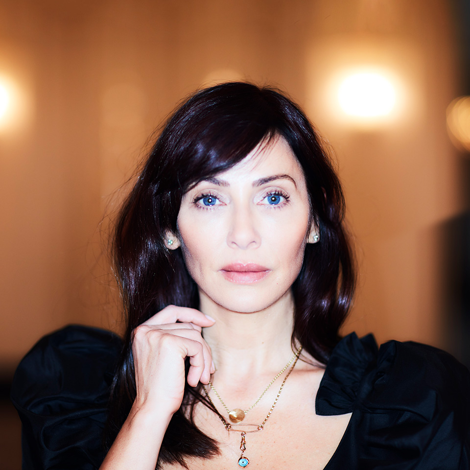 En kvinna med långt mörkt hår och blå ögon bär en svart dräkt och flera halsband och poserar inomhus mot en svagt upplyst bakgrund.