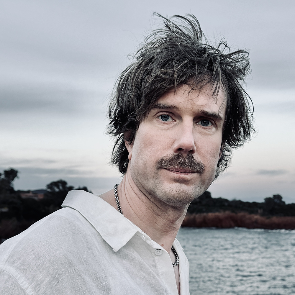 En man med rufsigt hår och mustasch i vit skjorta står utomhus vid ett vattendrag med träd i bakgrunden under en molnig himmel.