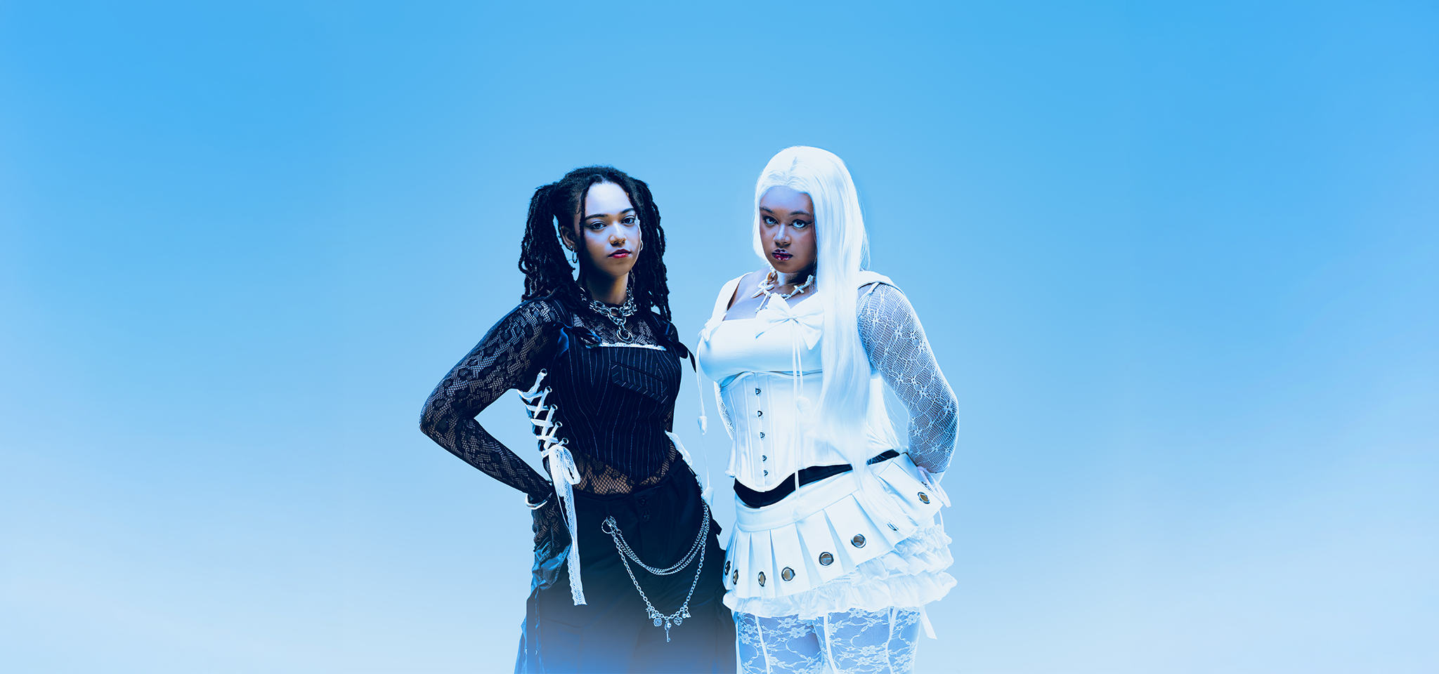 Två kvinnor står sida vid sida mot en blåtonad bakgrund; den ena bär svart spets och kedjor, den andra vit spets med korsett och långt vitt hår.