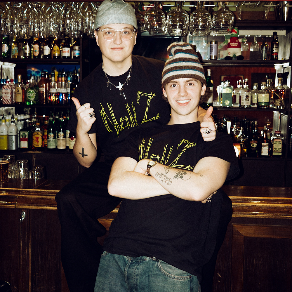 Två personer klädda i svarta grafiska t-shirts och hattar poserar tillsammans framför en bar med flaskor och glas.