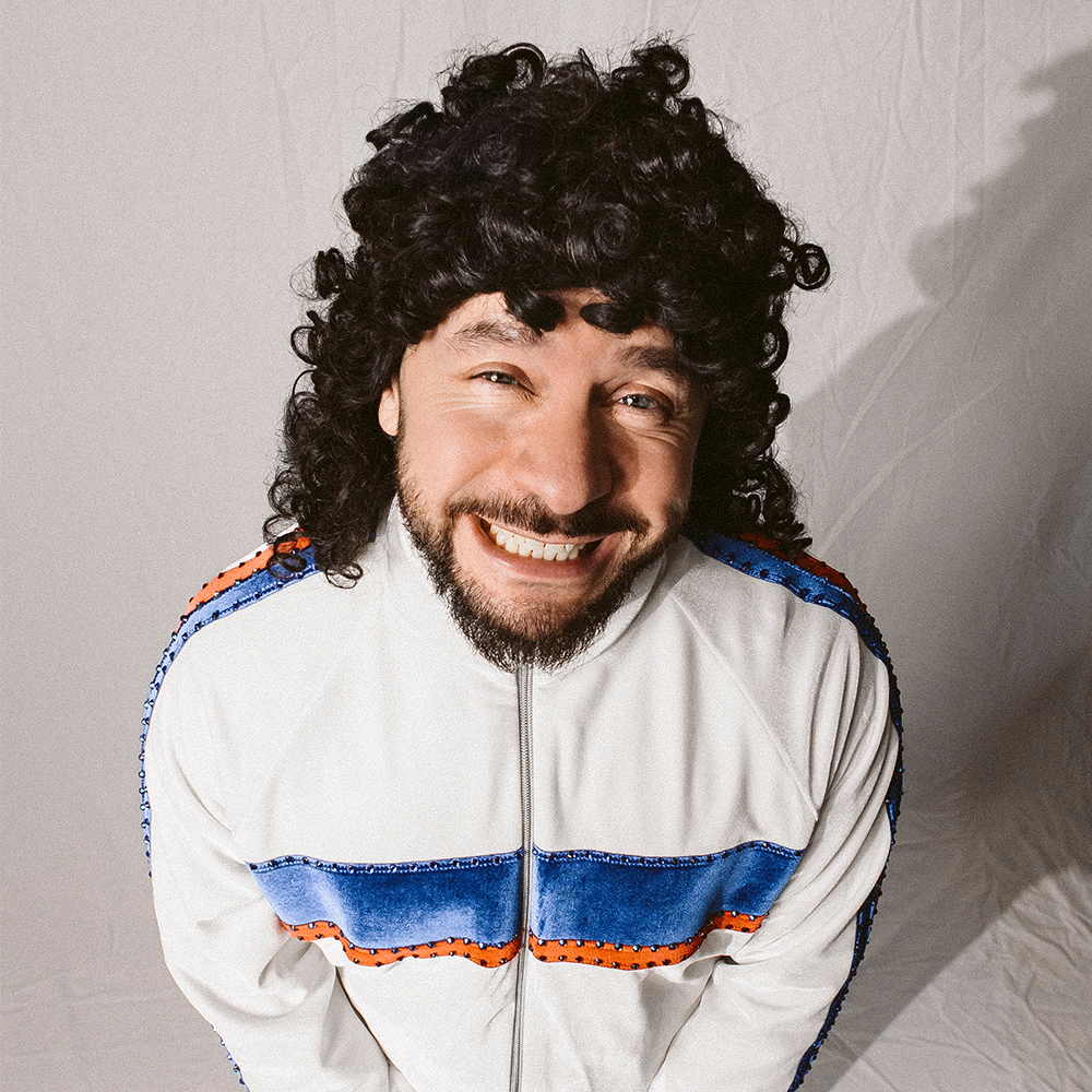 En man med lockigt mörkt hår och skägg ler brett iklädd en vit jacka med dragkedja och blå och orange ränder, stående framför en vanlig bakgrund.