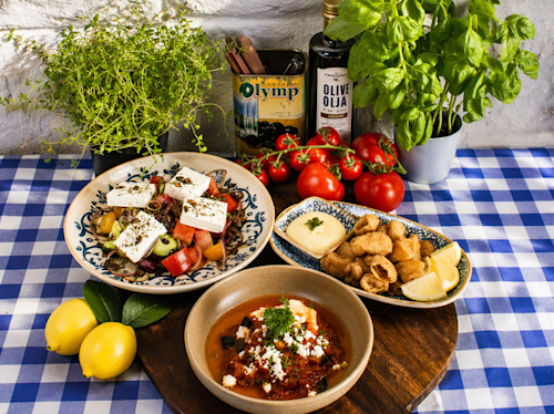 Three plates of Mediterranean food, fresh tomatoes, lemons, olive oil, herbs, and a tin of olives are arranged on a blue checkered tablecloth.