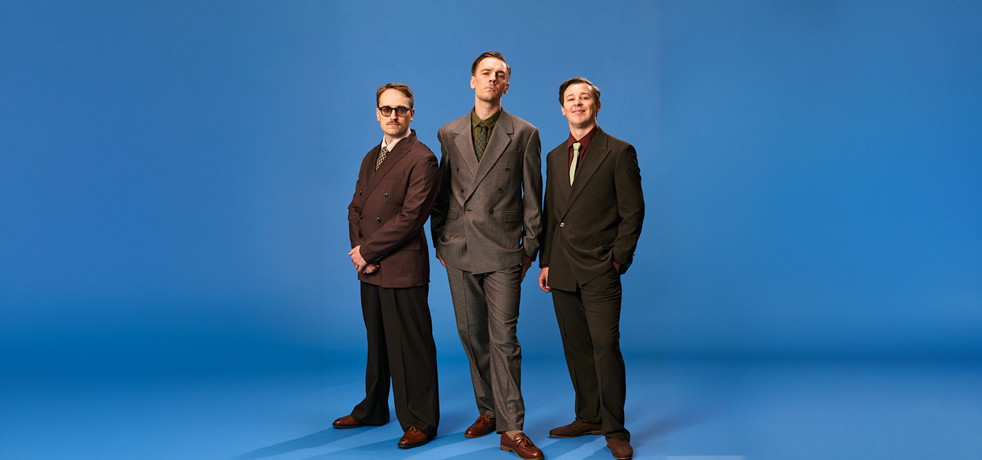 Tre kostymklädda män står sida vid sida mot en blå bakgrund, vända mot kameran med neutrala ansiktsuttryck.