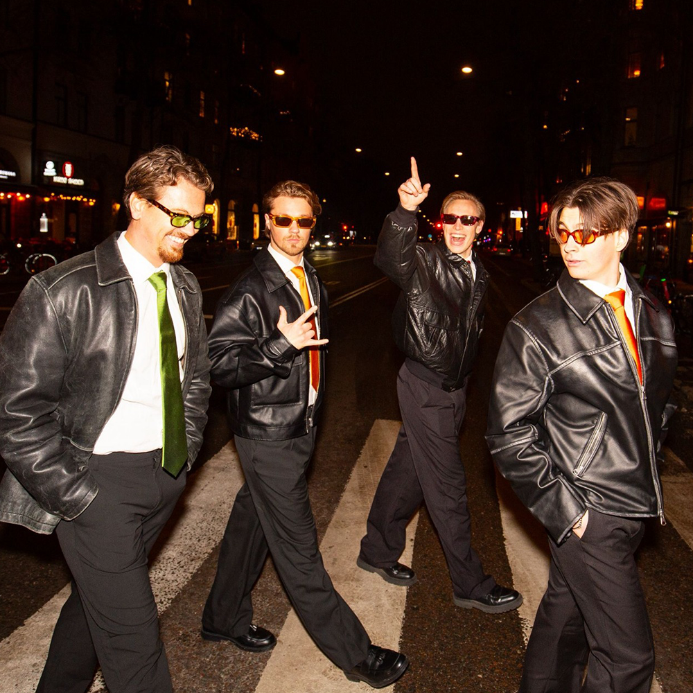 Fyra män i svarta skinnjackor och solglasögon går på ett övergångsställe på natten, med stadsljus och byggnader synliga i bakgrunden.