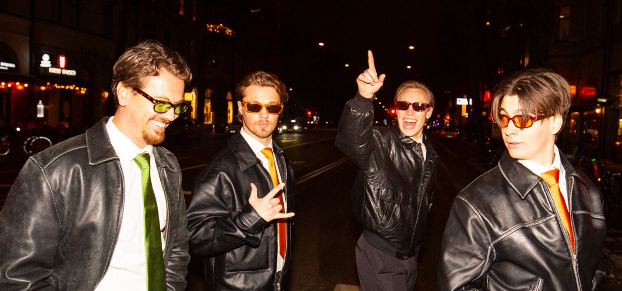 Fyra män i svarta skinnjackor, gröna slipsar och tonade solglasögon står på en gata i en stad på kvällen och poserar och ler.