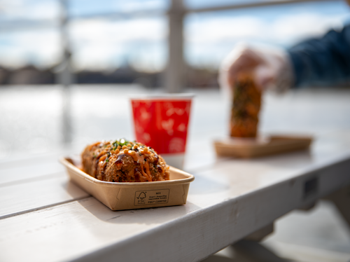 Close-up of a tray with five takoyaki balls topped with sauce and garnish on a white picnic table; a person picks up another tray in the background by the water.