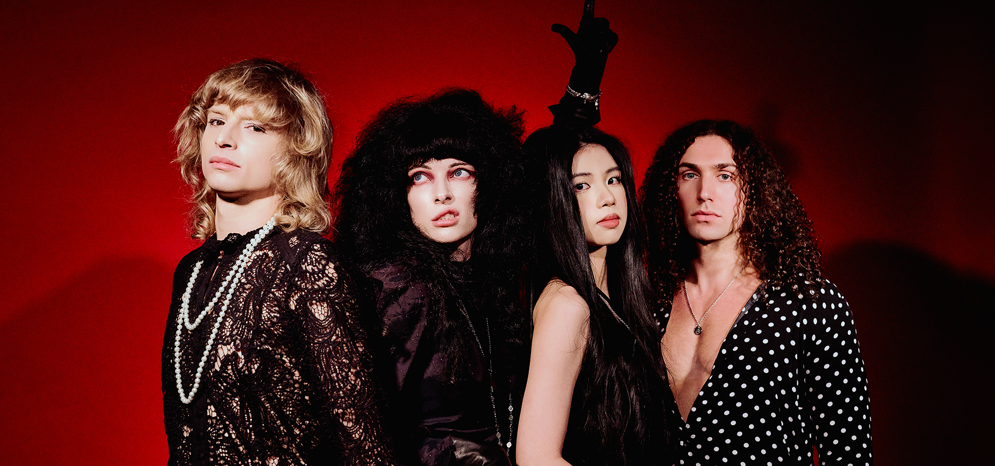 Fyra personer poserar mot en röd bakgrund, iklädda dramatiska svarta kläder och accessoarer, med varierande frisyrer och allvarliga ansiktsuttryck.