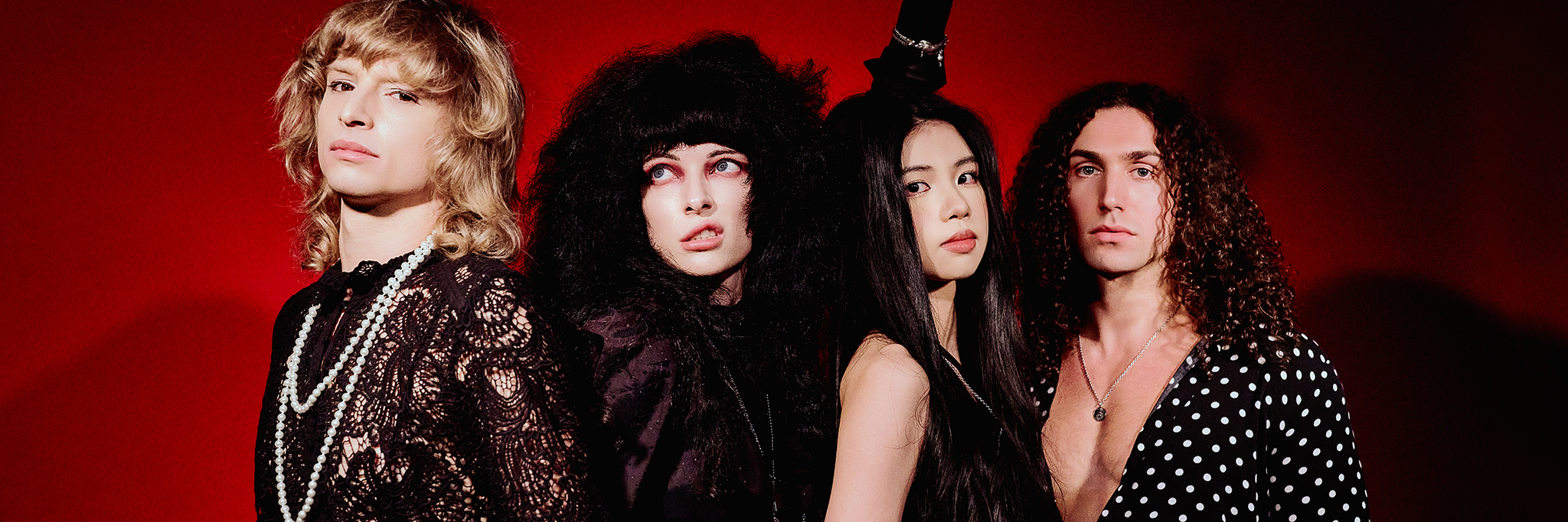 Fyra personer poserar mot en röd bakgrund, iklädda dramatiska svarta kläder och accessoarer, med varierande frisyrer och allvarliga ansiktsuttryck.
