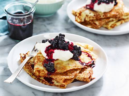 En tallrik med crepes toppade med vispgrädde och bärkompott, med en gaffel vilande vid sidan om. I bakgrunden syns en annan tallrik och en burk med bärsås.