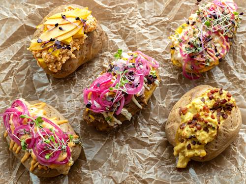 Five baked potatoes on crinkled parchment paper, each topped with various colorful ingredients like pickled onions, microgreens, and creamy sauces.