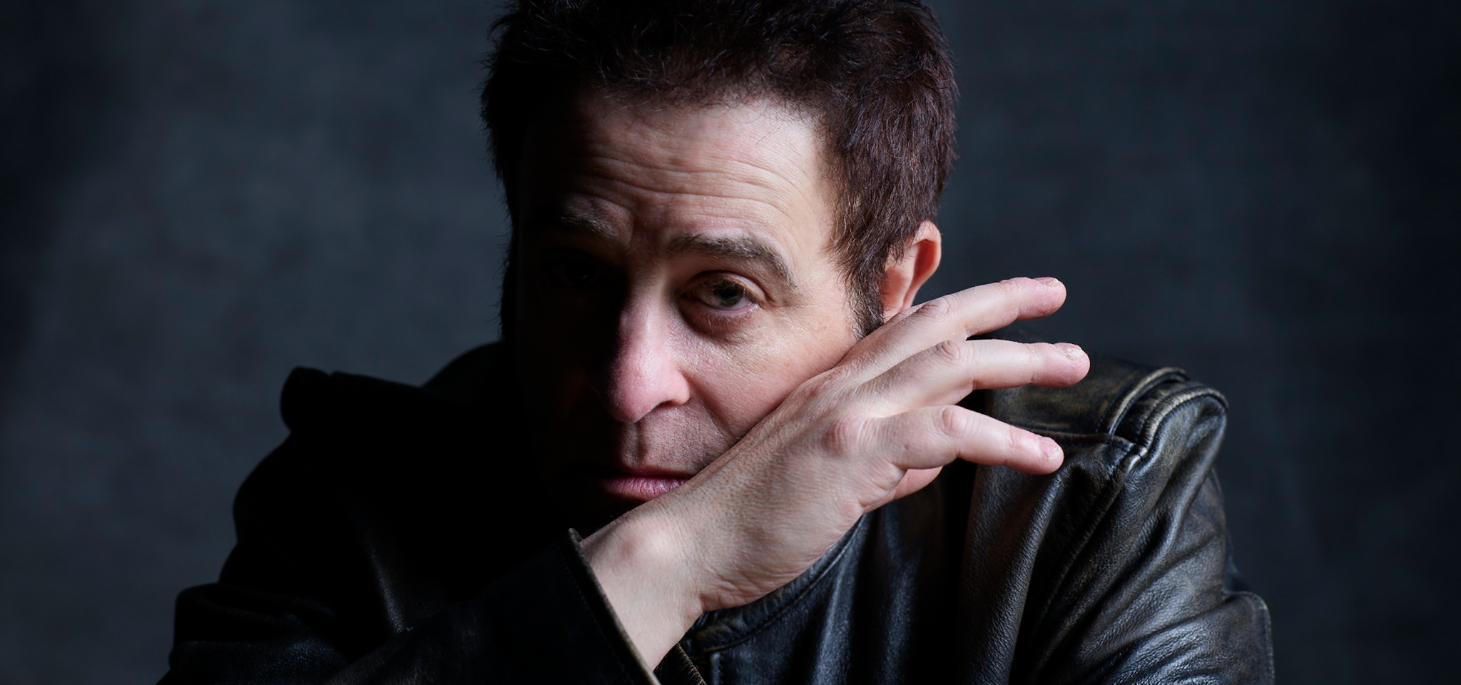 A person in a dark jacket poses with their hand partially covering their face against a dark background.