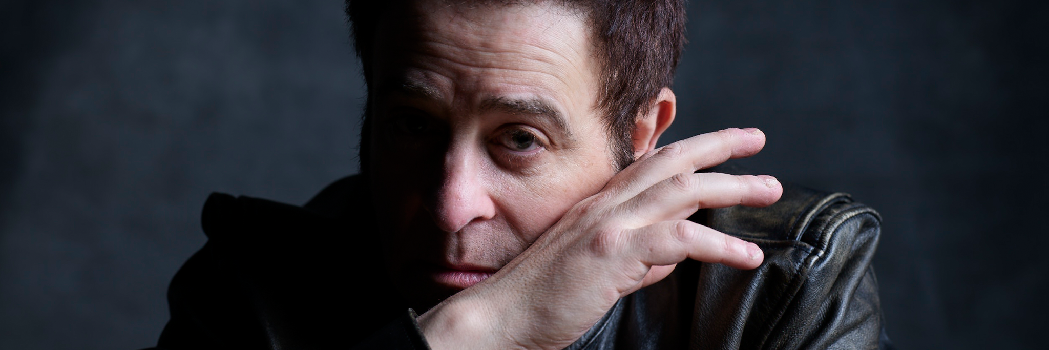 A person in a dark jacket poses with their hand partially covering their face against a dark background.