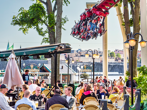 People sit at outdoor café tables while an overhead roller coaster passes by in the background near a body of water and city buildings.