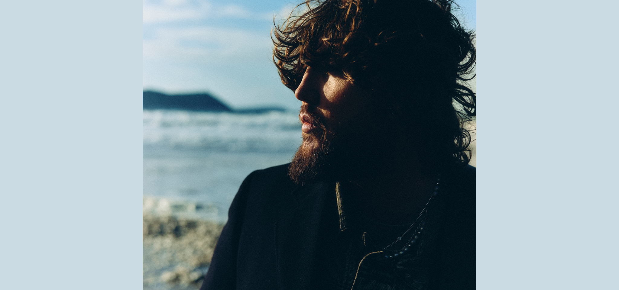 En man med lockigt hår och skägg står utomhus nära en strand, delvis i skugga, med havet och avlägsna kullar i bakgrunden.