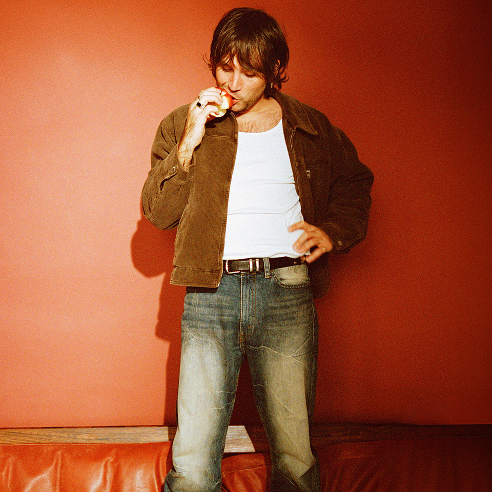 Man in a brown jacket and jeans stands against a red wall, looking down while eating an apple.