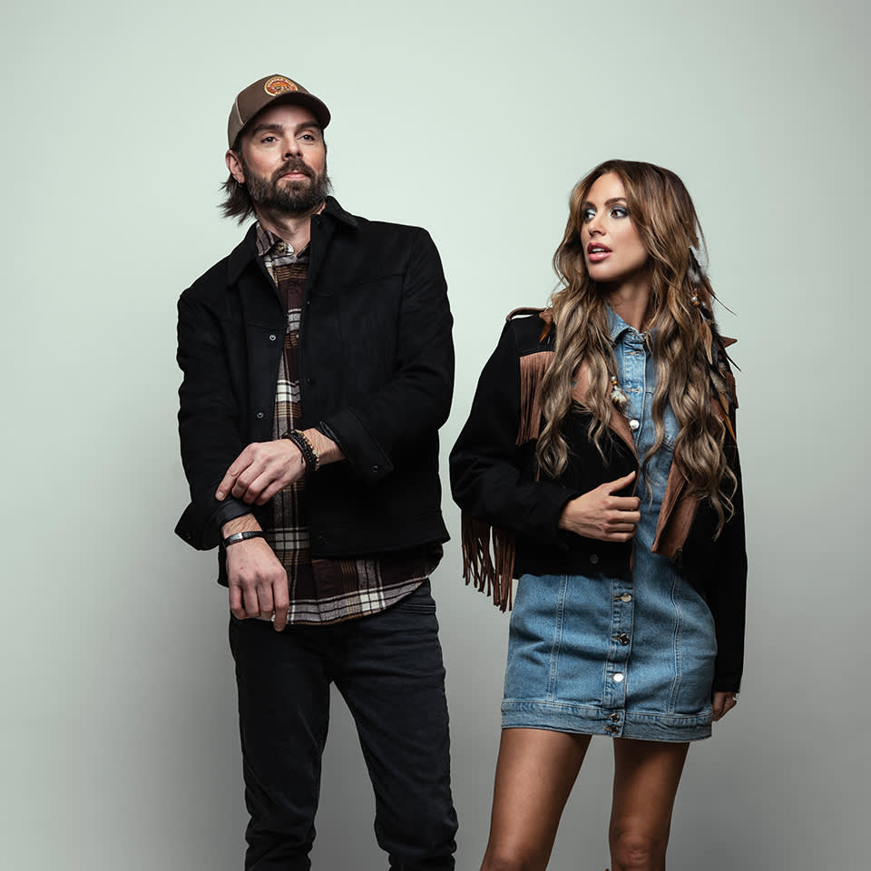 A man in a cap and plaid shirt stands next to a woman in a denim dress and fringe jacket, both posing against a plain light background.