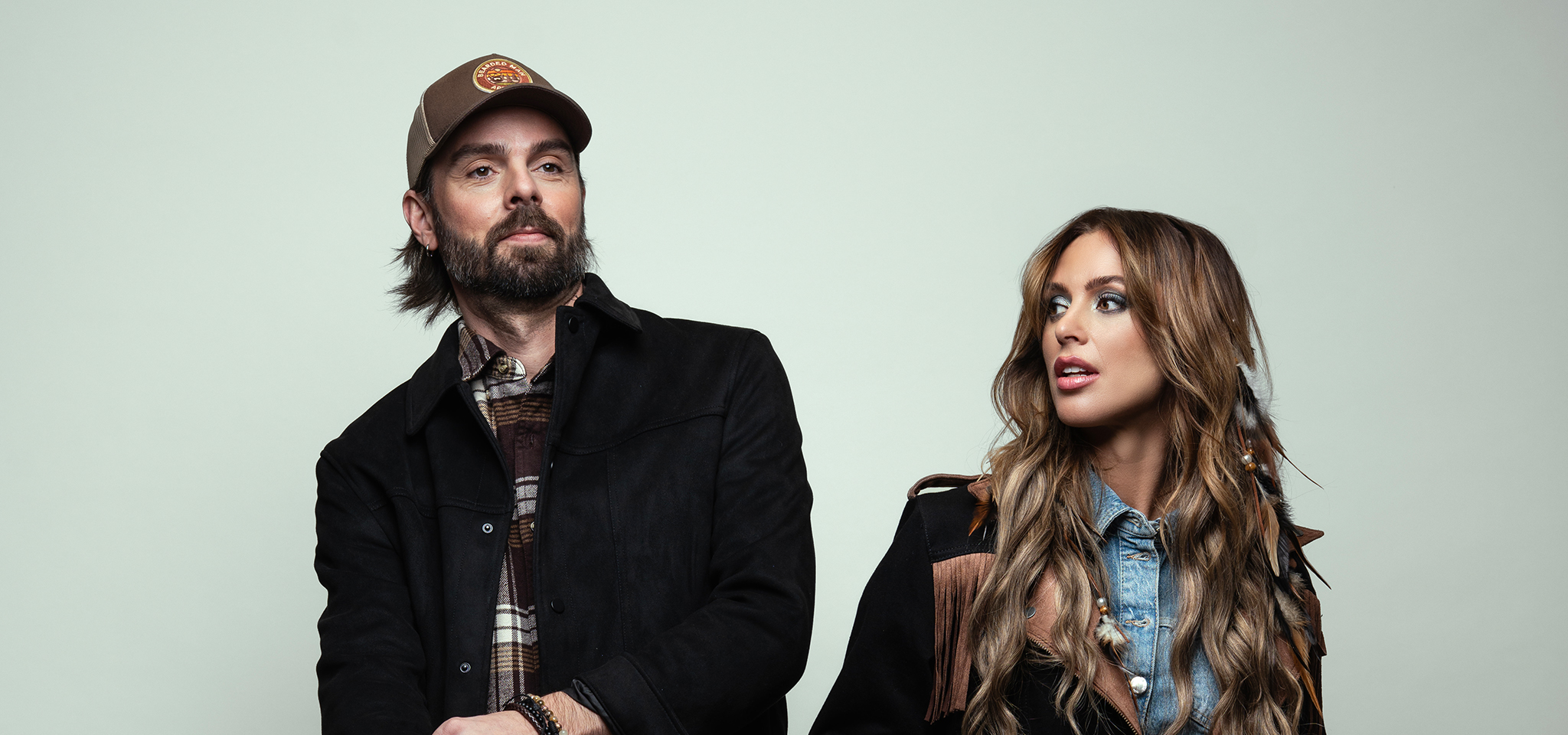A man in a brown cap and black jacket stands next to a woman with long wavy hair wearing a denim shirt and fringed jacket, both looking slightly to the side.