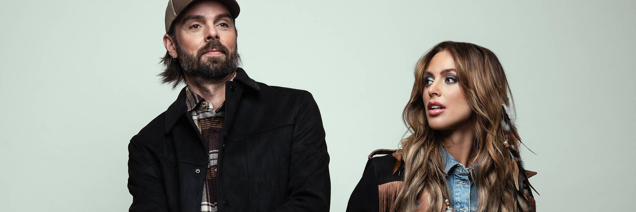 A man in a brown cap and black jacket stands next to a woman with long wavy hair wearing a denim shirt and fringed jacket, both looking slightly to the side.