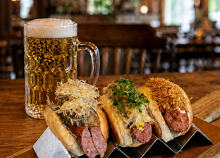 A glass of beer next to three gourmet sausages in buns, each topped with different garnishes, displayed on a wooden table in a rustic restaurant setting.