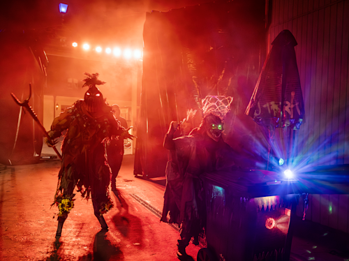Människor i kusliga dräkter med lysande ögon vandrar genom en dimmig, rödbelyst miljö på natten; en av dem skjuter en dekorerad vagn med starka lampor.