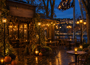 Outdoor café patio at dusk with string lights, pumpkins, and empty tables; trees with autumn leaves; roller coaster track and train overhead; city buildings visible in the background.