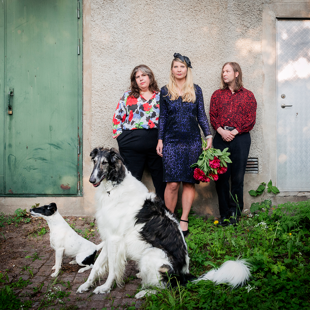 Tre personer står mot en strukturerad vägg, två med blommor i handen, och framför dem sitter två hundar på en gräsplätt med kullerstenar.