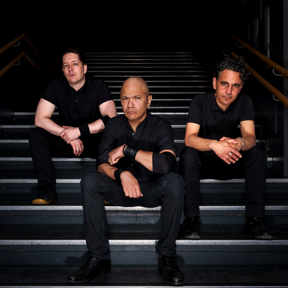 Three men dressed in black sit on a staircase, each looking directly at the camera in a dimly lit setting.