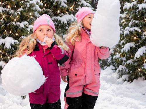 Två små barn i rosa vinterkläder som leker med snöbollar framför upplysta barrträd.