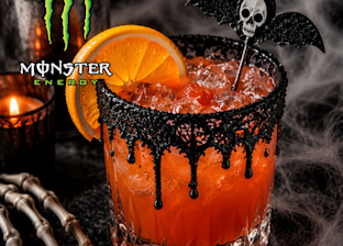 A glass of orange cocktail with black sugar rim, skull and bat stirrer, orange slice garnish, and Monster Energy logo. Candle, skeleton hand, and cobwebs in the background.