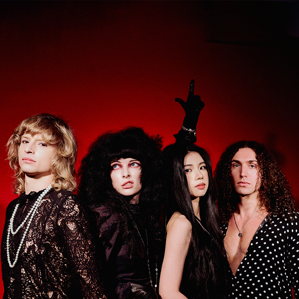 Fyra personer står framför en röd bakgrund, klädda i djärva, moderiktiga kläder och poserar med allvarliga ansiktsuttryck. En person höjer en hand med ett pekfinger.