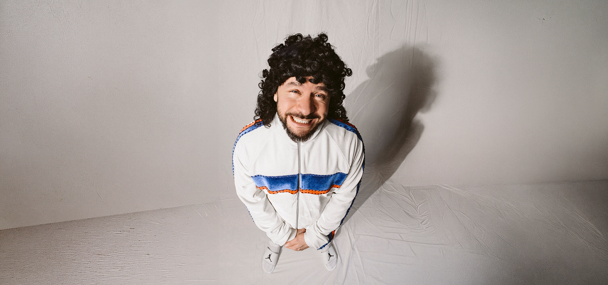 En man med lockigt hår och skägg står leende, iklädd en vit träningsoverall med blå och röda ränder, i ett enkelt, ljust rum.
