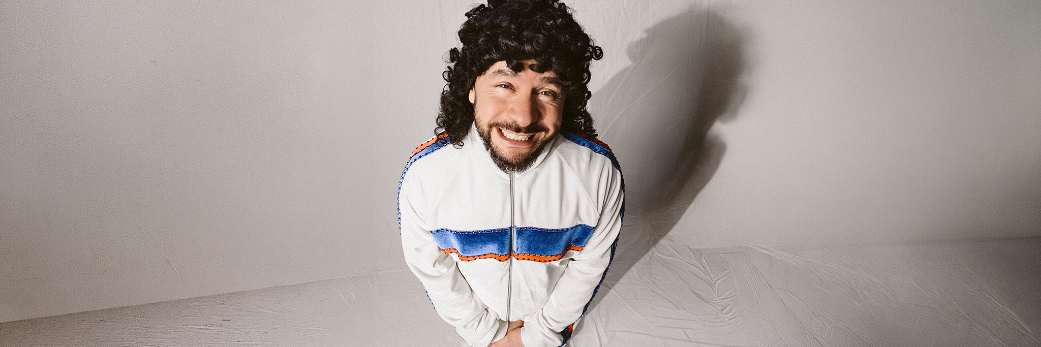En man med lockigt hår och skägg står leende, iklädd en vit träningsoverall med blå och röda ränder, i ett enkelt, ljust rum.