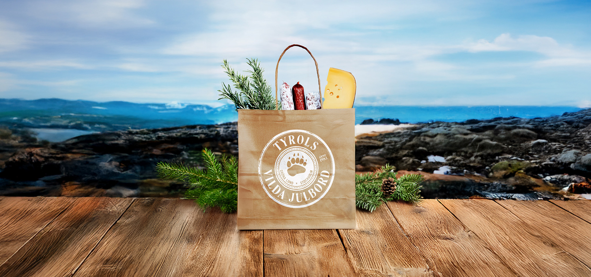 En papperspåse märkt "Tyrols Lilla Julbord" med ost, korv och örter ligger på en träyta med ett naturskönt landskap i bakgrunden.
