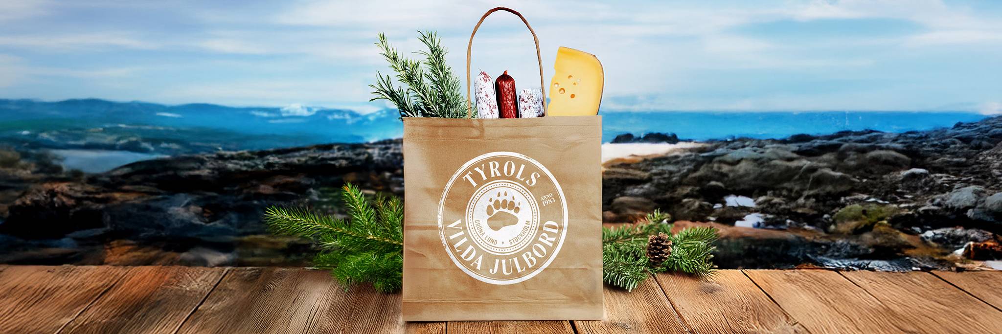 En papperspåse märkt "Tyrols Lilla Julbord" med ost, korv och örter ligger på en träyta med ett naturskönt landskap i bakgrunden.