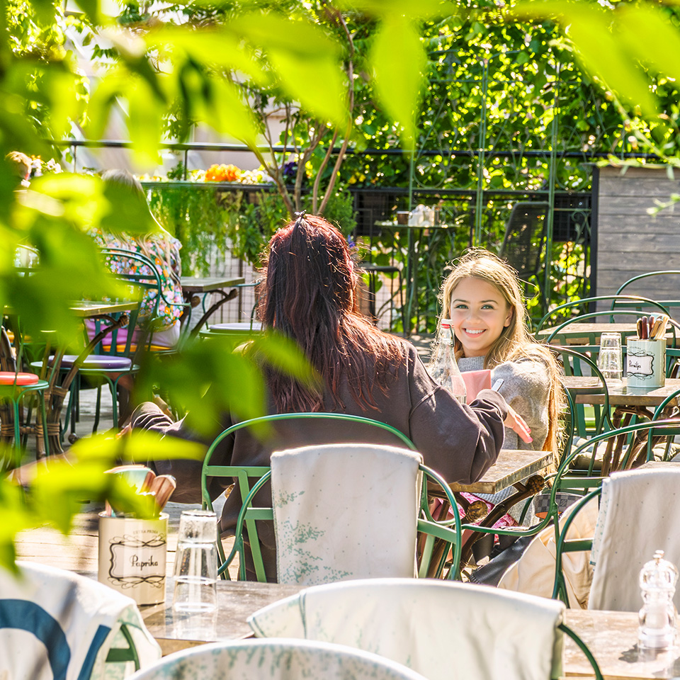 Two women sit at an outdoor café table on a sunny day, surrounded by green plants and empty chairs, with one woman smiling at the camera.
