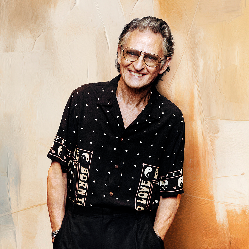 En medelålders man med grått hår och glasögon ler, klädd i en svartmönstrad kortärmad skjorta och mörka byxor, mot en beige och solbrun bakgrund.