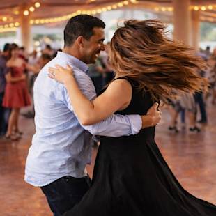 En man och en kvinna dansar tätt tillsammans under ljusslingor på ett utomhusevenemang, med andra människor dansande i bakgrunden.