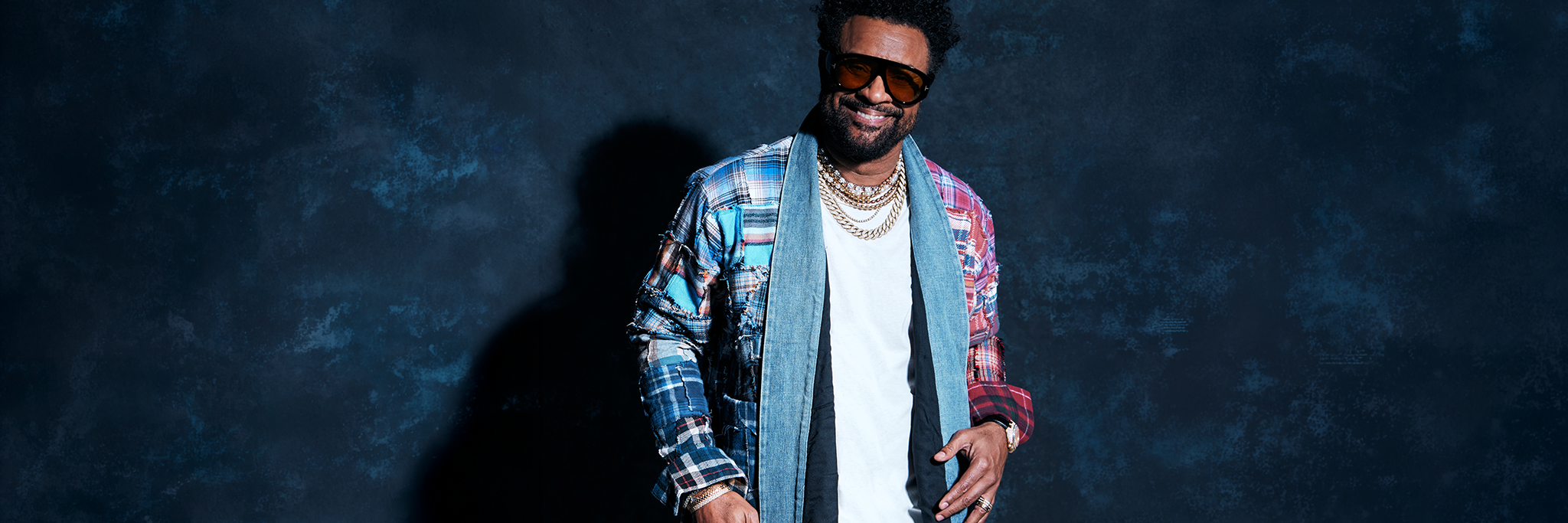 En man med solglasögon, halsband i flera lager, en flerfärgad jacka och jeans står och ler mot en mörk bakgrund.