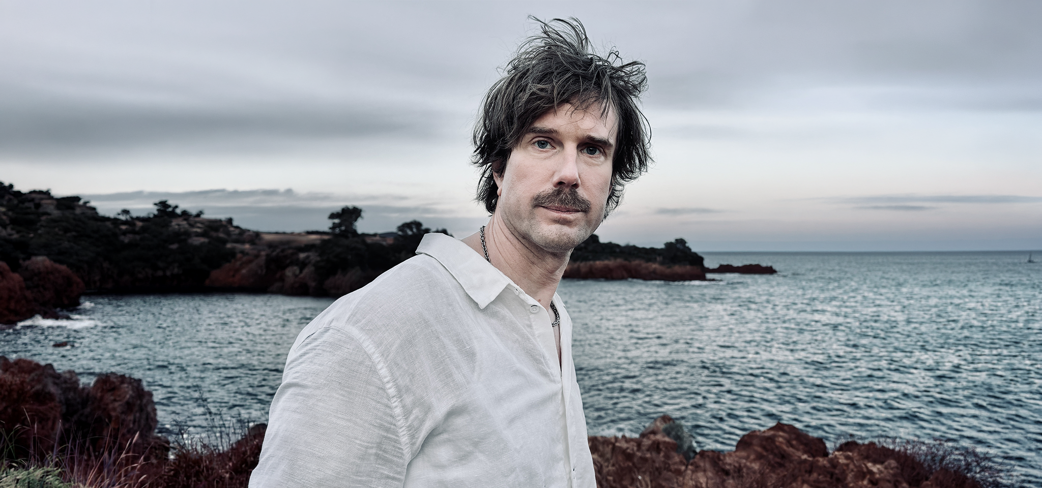 En man med rufsigt hår och mustasch, klädd i ljus skjorta, står vid en klippig kustlinje med havet och molnig himmel i bakgrunden.