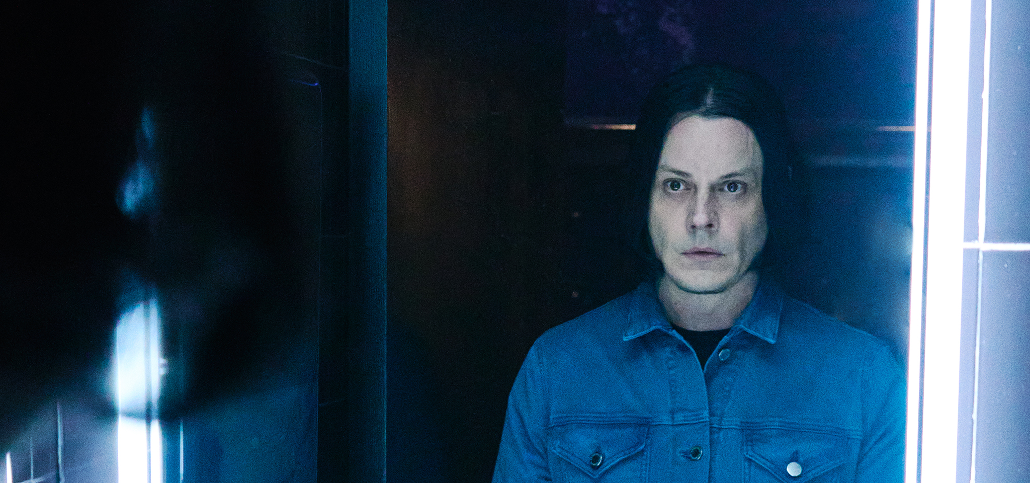 A person with shoulder-length dark hair and a blue jacket stands in front of a mirror, illuminated by vertical white lights.