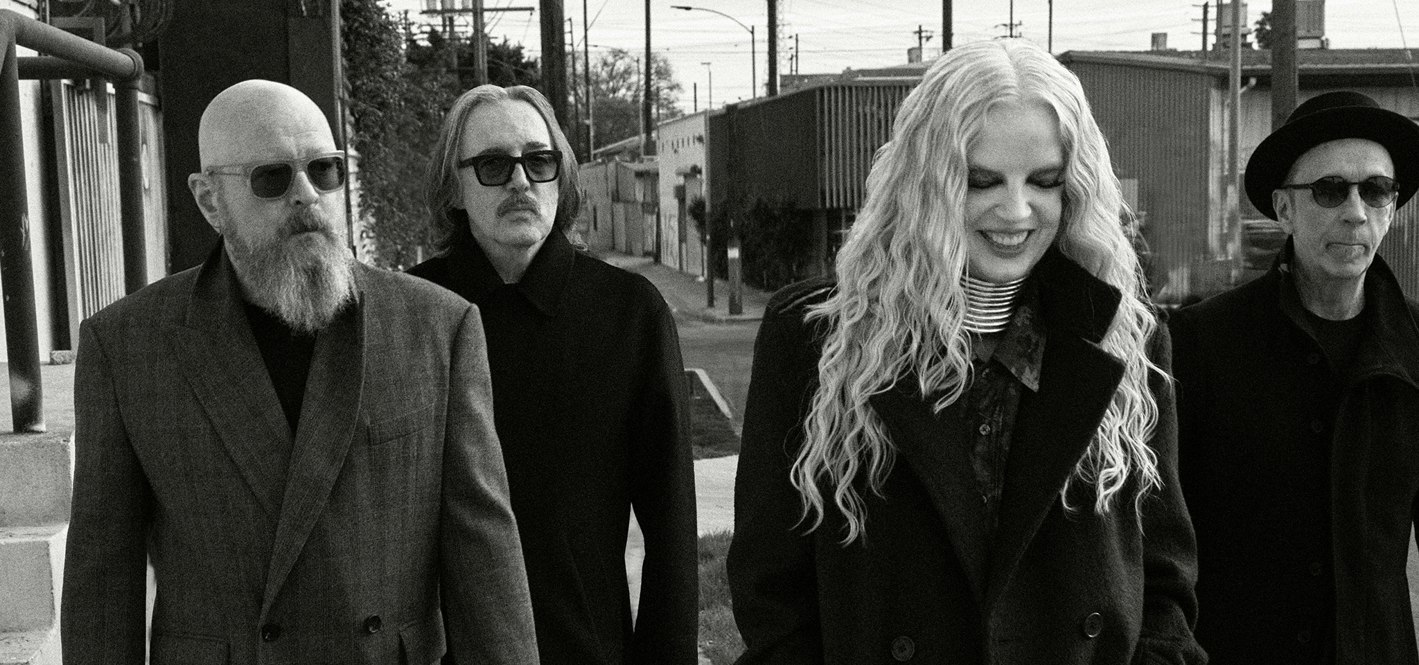 Fyra personer i mörka rockar går utomhus i en stadsmiljö; en kvinna med långt hår ler medan de andra tittar rakt fram.