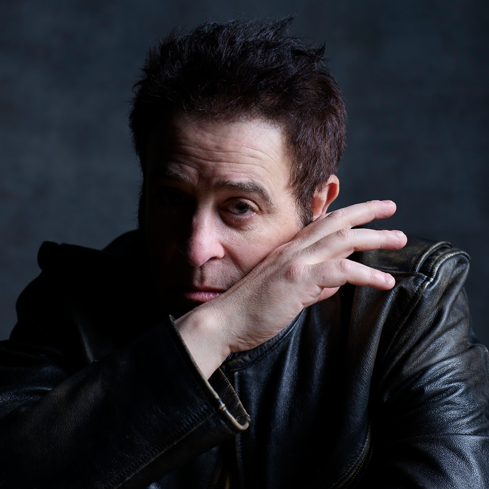 A person in a dark leather jacket sits against a dark background, resting their hand across their face and looking toward the camera.
