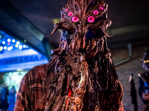 Person iförd en detaljerad, utomjordisk mask med glödande rosa ögon spelar saxofon inomhus i artificiell belysning.