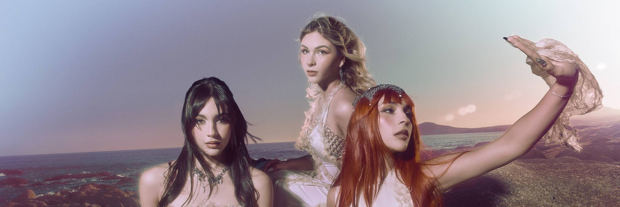 Three women in white dresses pose on a rocky beach at sunset, with one raising her arm and the ocean visible in the background.