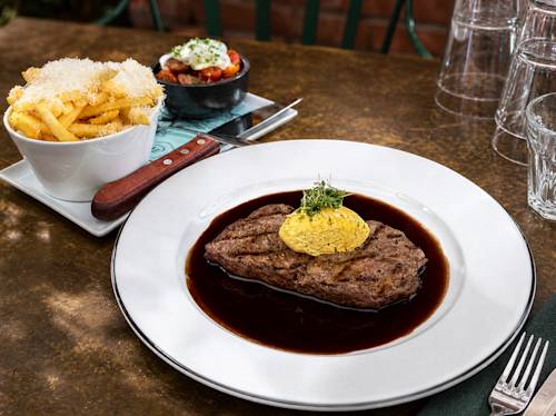 A steak with herb butter sits in a dark sauce on a white plate, served with a bowl of seasoned fries and a small bowl of vegetables on a brown table.