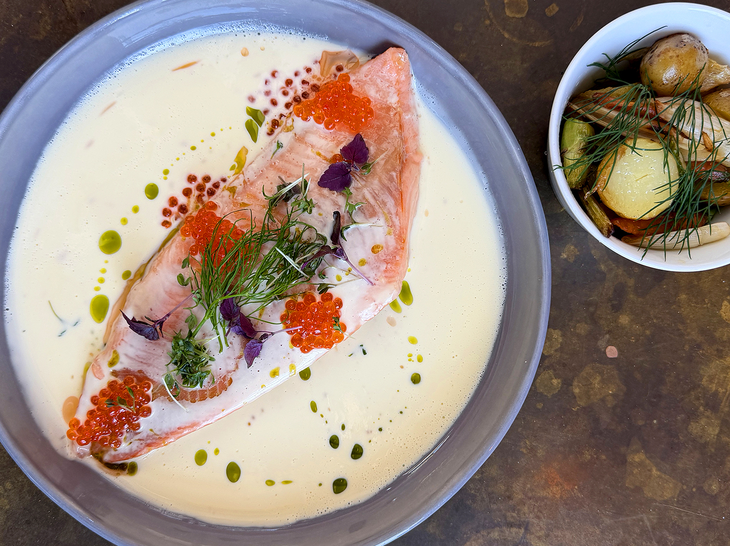 En tallrik med pocherad lax i en krämig vit sås, garnerad med fiskrom, örter och olja, bredvid en skål med ugnsbakade potatisar med färsk dill.